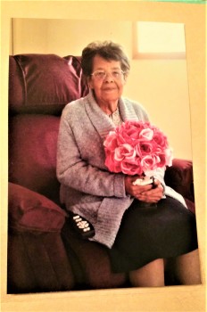 Esther R sitting in her lift chair. She is smiling and holding a bouquet of pink flowers. She has short dark hair and is wearing a lilac cardigan with a calf length black skirt.