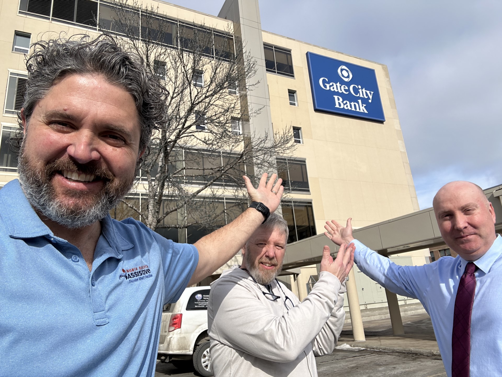 Tom Gerhardt and Mike Chaussee of North Dakota Assistive join Kevin Warner of Gate City Bank in Fargo outside of the downtown office for a photo. The Gate City Bank logo is in the back on the outside of the building, and Tom, Mike, and Kevin are all gesturing toward it with their arms in the air.