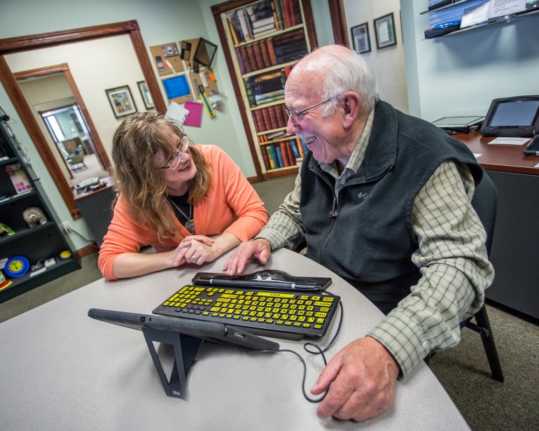 Sr. AT Consultant Jeanne Krull sits with an older male who is learning how to use a tablet to zoom into pictures.