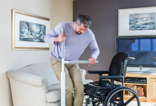 Picture of man moving from rising from the couch using the Advantage Rail. One hand holds the vertical bar of the rail and the other holds the horizontal rail. He is transferring from the couch to the wheelchair.