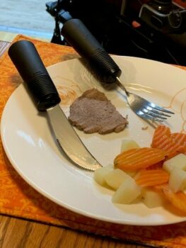 image shows an adaptive knife and fork on a plate with meat, potatoes and carrots. The silverware has enlarged and weighted handles which are black in color.