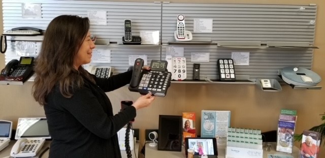 Roxanne is in front of a wall of specialized phones - there are about a dozen of them on a wall and counter display. She's holding a phone and looking at it. The phone is black with large buttons and a text display.