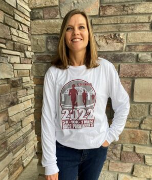 woman standing outside near brick wall wearing 2022 Race for Parkinson's fundraiser shirt that is white with maroon and grey logo with runners 