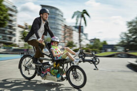 Man wearing suit jacket and helmut is giving a bike ride to younger girl wearing helmet in a sunny city on a tandem bicycle