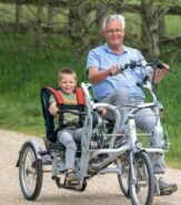Older smiling man in blue shirt is riding a double seated adapted bicycle with a smiling little boy harnessed in beside him.