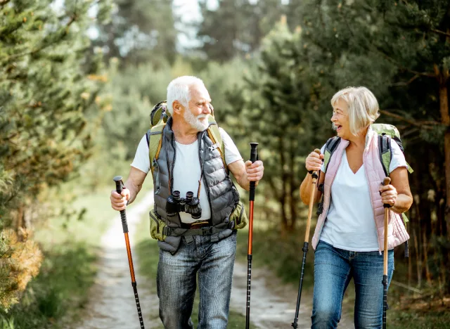 Older man with grey hair and beard wearing white shirt and vest using walking sticks outside is smiling and walking with older lady with grey hair wearing white shirt and pink vest using walking sticks. They are both walking outside on a trail with trees around them.