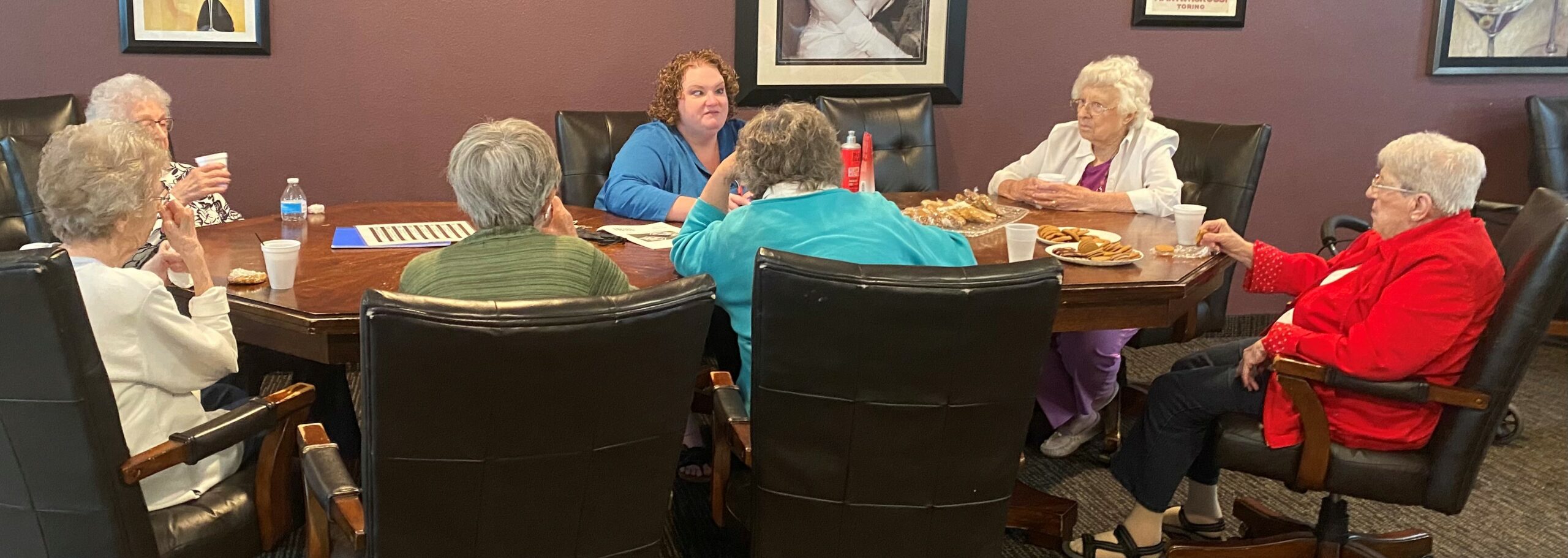 Seven people around an oval table. The woman facing the camera on the far end of the table leads the conversation. The others look a her attentively.