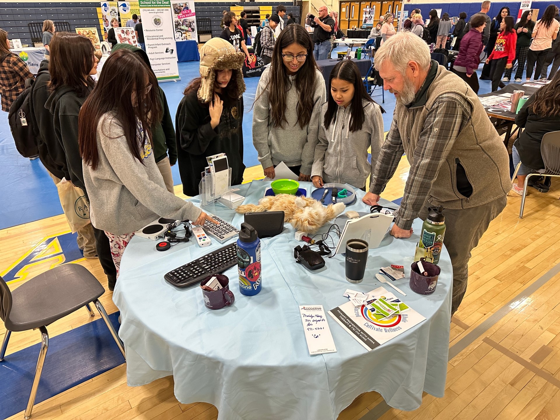 ND Assistive Executive Director Mike Chaussee leans over a circlular table at a tabling event at Standing Rock High School in Ft. Yates. A group of students are gathered around listening to Mike explain assistive technology items on display.