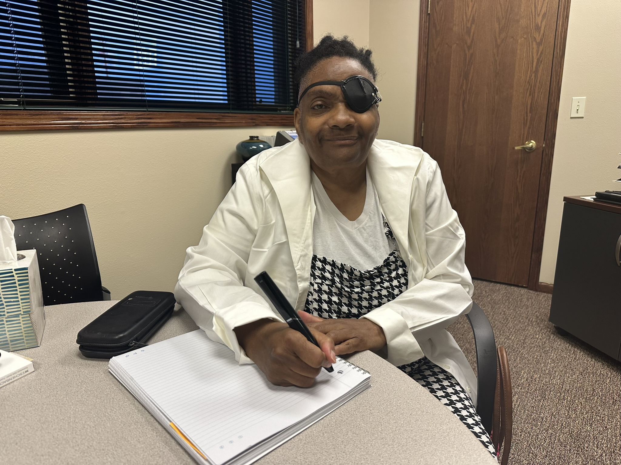 Rita Plair is pictured with a smart pen she received through North Dakota Assistive. Rita is wearing a white coat with a white t-shirt underneath and a black and white checkered dress.