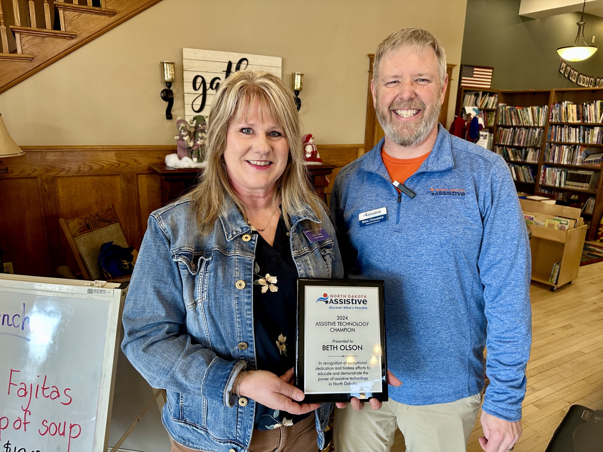 Beth Olson holds a plaque honoring her as a 2024 Assistive Technology Awareness Champion. The award was presented by executive director Mike Chaussee, who is standing by her side.
